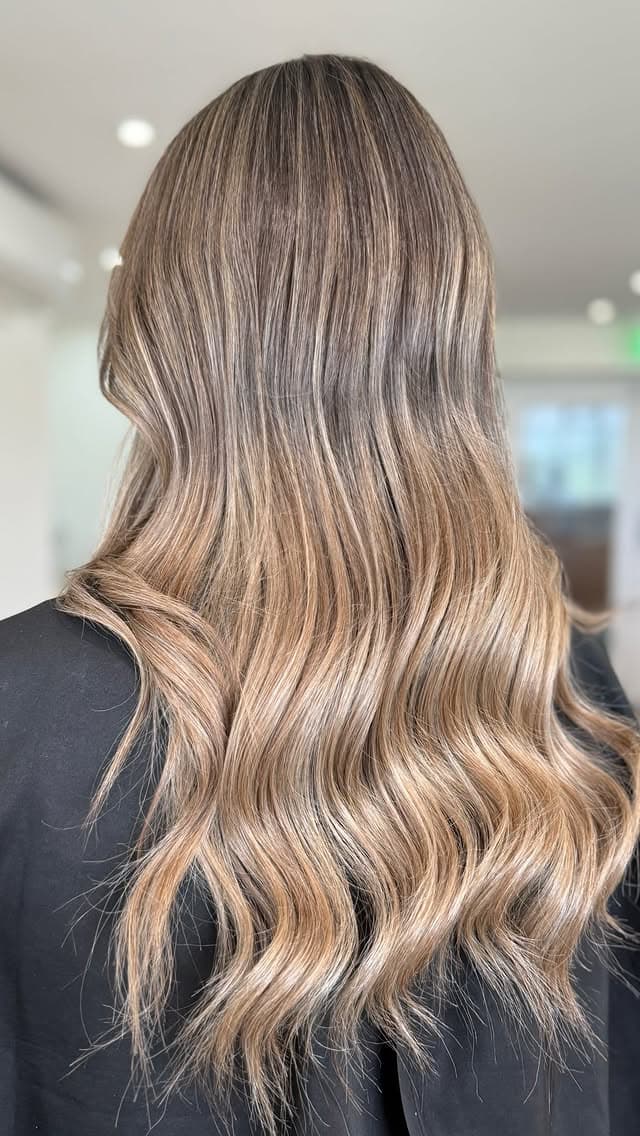 A brunette root melting to warm-bronde waves in the bright warm-concrete salon interior.
